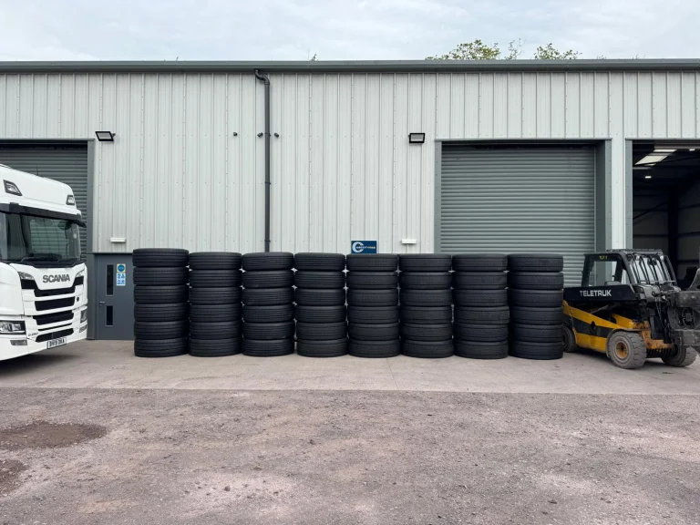 Rows of black remould HGV tyres are neatly stacked outside a modern industrial warehouse in Shropshire. A white Scania lorry is partially visible on the left, while a yellow forklift is parked on the right beside the tyre stacks. Roller shutter doors and a clean concrete yard create a professional commercial setting. These high-quality remould HGV tyres are available in Shropshire for commercial vehicle fleets and operators.