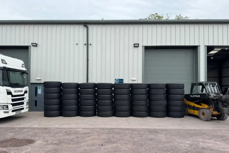 Rows of black remould HGV tyres are neatly stacked outside a modern industrial warehouse in Shropshire. A white Scania lorry is partially visible on the left, while a yellow forklift is parked on the right beside the tyre stacks. Roller shutter doors and a clean concrete yard create a professional commercial setting. These high-quality remould HGV tyres are available in Shropshire for commercial vehicle fleets and operators.